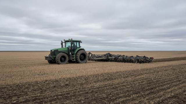 A green tractor pulling a seeder across a harvested field under a gray sky.