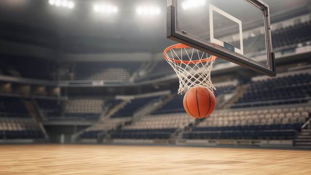 A basketball dropping through the net below a glass backboard in a brightly lit arena with empty seats